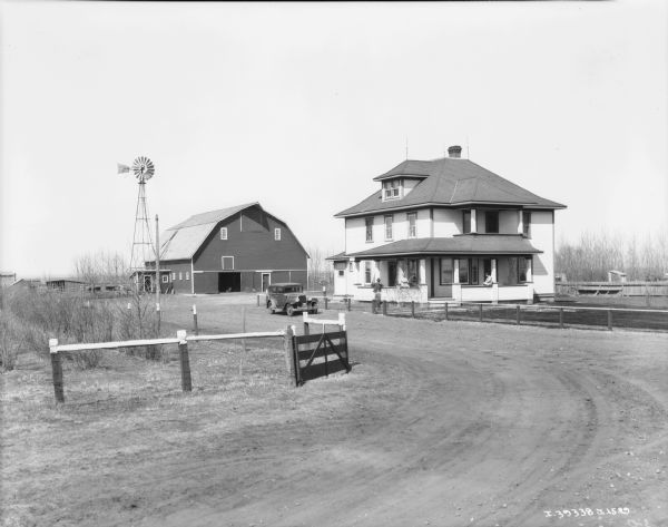 View down curving road towards a family posing on the porch of a farmhouse. There is an automobile parked near the house, and there is a fence, farm buildings and a windmill in the farmyard.