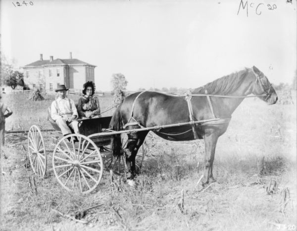 A man and woman are sitting in a horse-drawn wagon in a field. In the background is a farmhouse. Another man is standing on the far left.