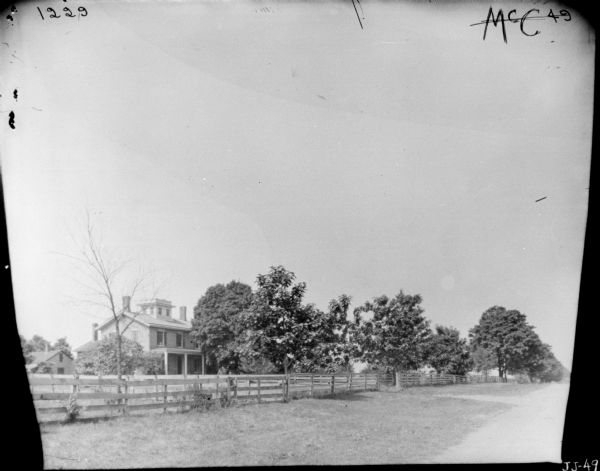 View from road towards a two-story farmhouse. The house has a front porch, and on the roof is a square cupola, or belvedere, with two windows on each side. A long wooden fence runs along the road in front of the fields and lawn of the property. There are outbuildings in the background.