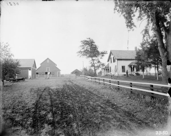 View along fence towards a barnyard on the left, and a man and two women on the porch and in the yard in front of a farmhouse on the right. The man is standing and is holding on to what may be a push mower.