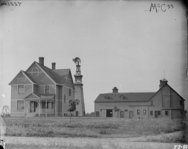 View across road towards a large farmhouse. There is a windmill just behind the house, with a tall wooden enclosed structure at the base. A barn is on the right. A lawn swing is on the left side of the house. A few small new trees have been planted in the lawn.