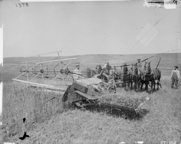 A group of three men, and a child, are posing on and around a team of six horses powering a McCormick push binder in a field. In the distance are farm buildings and low hills.