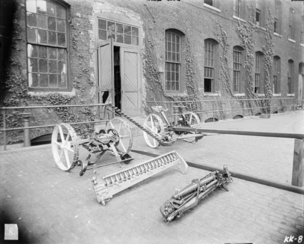 Mowers set up outdoors in front of a brick factory building. One of the mowers is assembled, and one mower is packed in separated pieces ready for shipping.