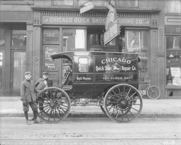 View of two teenage boys posing with an International Highwheeler in front of the storefront of the Chicago Quick Shoe Repair Co. 120 James Street.
