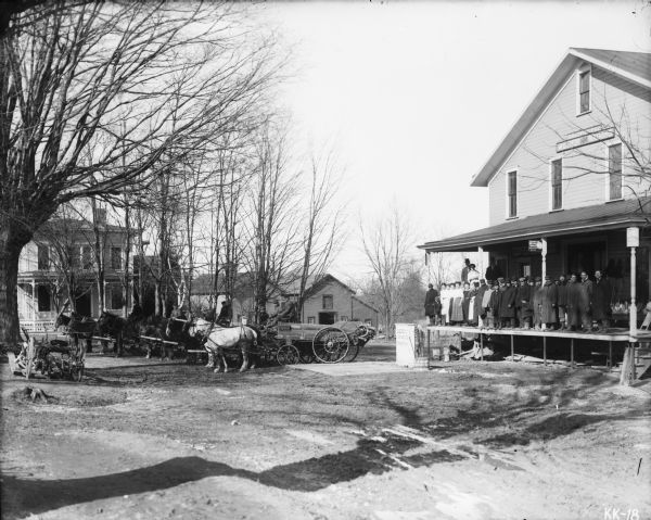 View towards a large crowd on the porch of a dealership. There is a sign on the ground in front of the building, perhaps over a scale, that reads: "Osgood US Standard, Binghamton, N.Y." A sign above the porch of the building reads: "D.D. Costello Co., Weber Wagons." There are men driving horse-drawn manure spreaders in the yard.