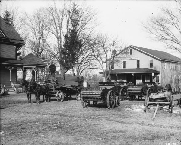 View towards a man standing on a horse-drawn manure spreader on the left. In the background is a dealership, with a sign above the porch of the building the reads: "D.D. Costello Co., Weber Wagons."