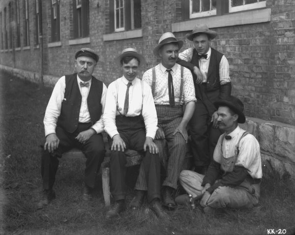 Five men in "picnic clothes" are posing on the grass alongside a brick building. They are all wearing neckties or bow ties. The man sitting in the grass on the right is holding a hammer.