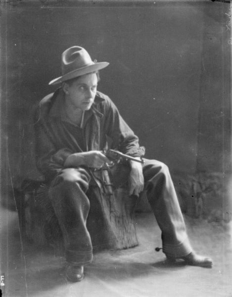 Studio portrait of a man wearing a hat, shirt, pants with fringe along the sides, and spurs on his boots, sitting and holding a gun.