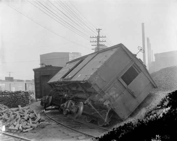 View towards a boxcar tipped over on railroad tracks. The boxcar is leaning on a large pile of what may be gravel. Piles and stacks of wood are on the left, with a row of boxcars in the background.