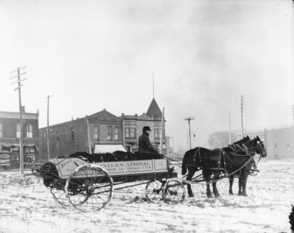 View towards a man driving a horse-drawn manure spreader in a field in a residential area, (test plot?). A sign on the side of the manure spreader reads: "Manure Spreader No. 3." There are storefronts in the background.