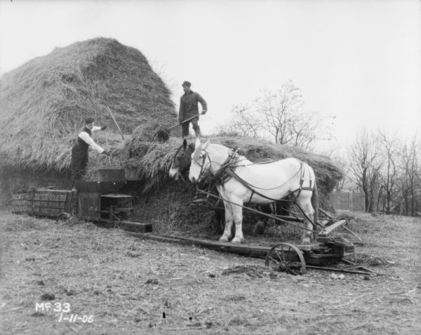 Men are feeding hay into a hay press from large stack. A team of horses is powering the hay press.
