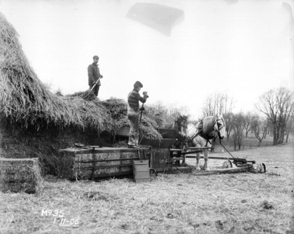 Men are feeding hay into a hay press from large stack. A team of horses is powering the hay press.
