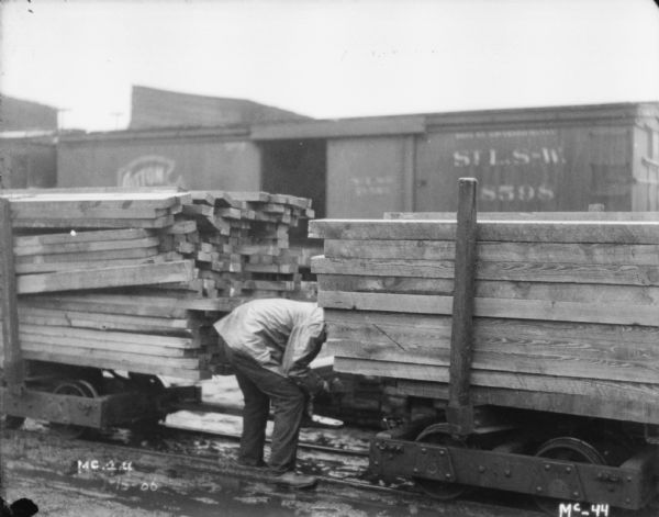 View towards a man hitching flatcars loaded with lumber. Railroad cars are in the background.