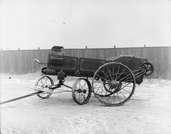 Left side view of a manure spreader parked in a yard at McCormick Works. There is a fence in the background.