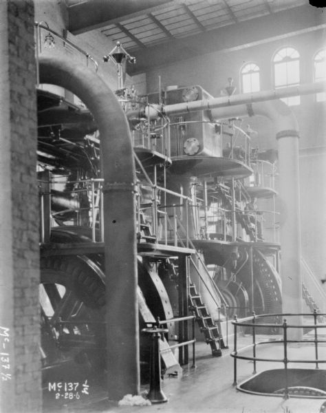 Turbines inside plant at McCormick Works. A man is sitting on a set of steps on a platform above the floor.