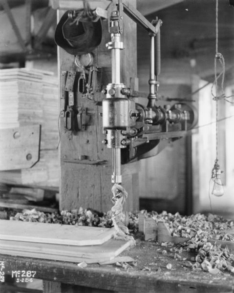 Drill inside McCormick Works. Wood shavings are on the bench, and a hat is handing from a post behind the drill. A light bulb is hanging from a wire on the right.