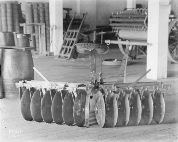 Disk harrow indoors. Barrels and parts are in the background.