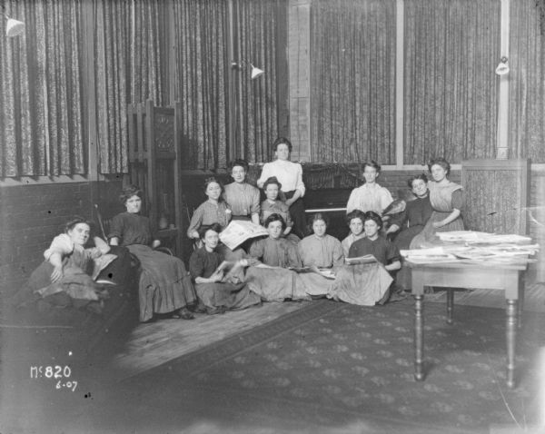 Formal group of women relaxing in the corner of a room. There is a large clock against the wall on the right, and a piano in the corner. One woman is lying on a lounge on the far left, and a group of women sitting together are holding newspapers. A stack of other papers are on a table on the right.