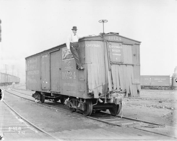 A man wearing a hat, bow tie and suspenders is posing standing on a ladder on the side of a boxcar at McCormick Works. There is a poster on the side that reads: "Deering Threshing Machines." The front wall of the boxcar is burst out. Perhaps from an accident or contest.