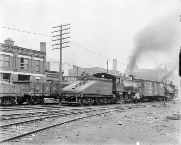 Illinois Northern rail road train, with two engines, a coal car and a car with front wall burst out, at McCormick Works. There are commercial buildings in the background.