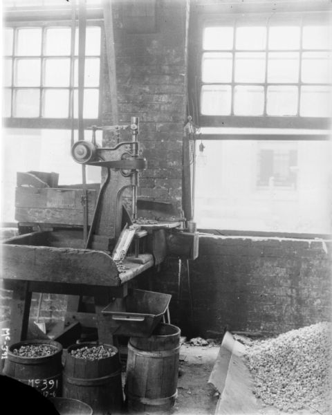 View of a drill in a work area near two large, open windows. A large pile of metal nuts are on the floor on the right, and more metal nuts are in barrels under the drill.