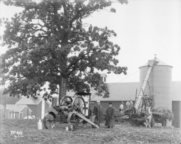 A group of people are posing on a farm where a tractor is powering a thresher. Men and boys are standing on and near a horse-drawn wagon and a corn elevator. They are filling a silo next to a barn. Vicinity of Chicago.