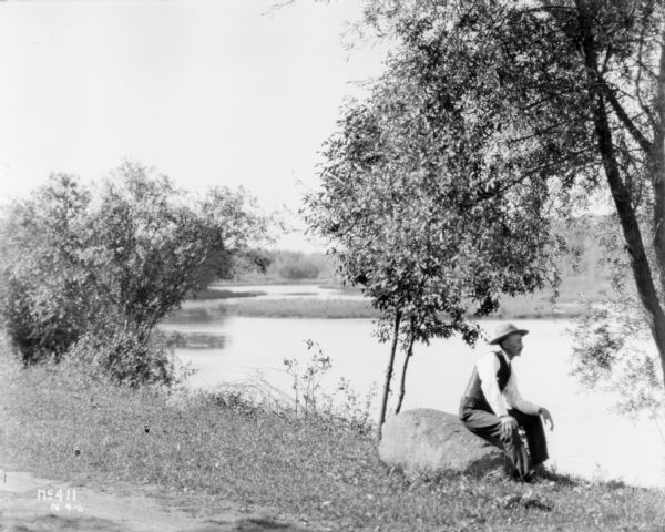 A man is sitting on a rock by a river's edge under trees. Vicinity of Chicago.