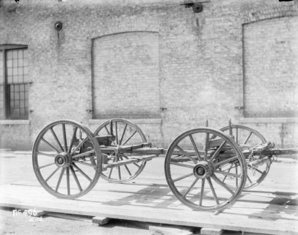 View of a wagon, without wagon box, set up on a wooden pallet outdoors. A brick factory building is in the background.