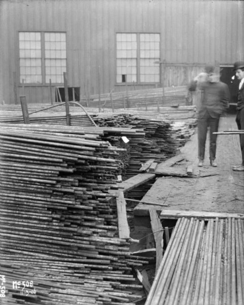 Two men are standing on a wooden loading dock near the pipe storage area. A factory building is in the background.