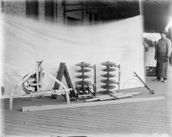 Disk harrows dismantled, ready for crating at McCormick Works. There is a light-colored backdrop set up behind harrows, and a man is standing on the right.

