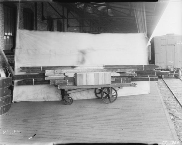 Hay stackers crated for delivery on a loading dock at McCormick Works, with a light-colored backdrop set up in the background. There is a railroad car on the railroad tracks on the right.