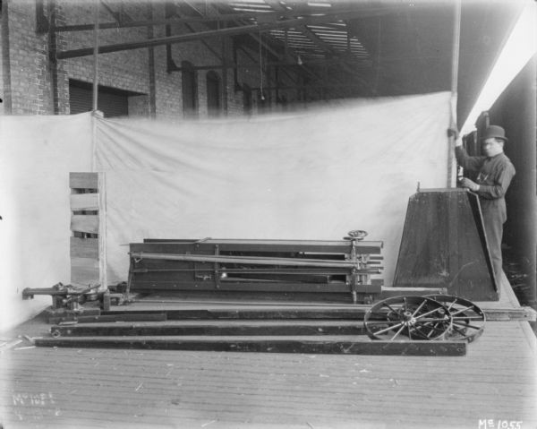 Hay stackers crated for delivery sitting on a loading dock at McCormick Works. There is a light-colored backdrop set up in the background. A man is standing near the backdrop on the right.
