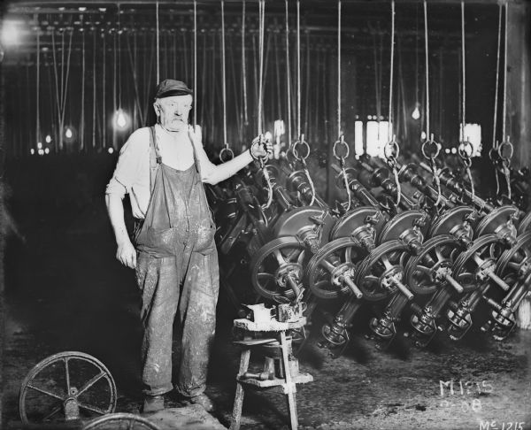 McCormick Works employee Peter Butzen with paint can and brush, applying pin-stripes to binder parts. He is posing with mower drivetrains hanging from hooks inside IHC's McCormick Works paint and pinstriping room, Chicago, Illinois. There are details of mower pin-striping. In operation from 1873-1961, the works was located at Blue Island Avenue and Western Avenue in the Chicago subdivision called "Canalport." The works encompassed 160 acres of multi-story buildings and 103.3 land acres at maximum. It later closed in 1961 due to technological and architectural obsolescence. Products: binders, reapers, headers, push binders, corn machines, harvester-threshers, mowers, rakes, sweep rakes, hay stackers, huskers and shredders, ensilage cutters, knife cutters, cotton machines, seeders, farm trucks, fertilizer distributors, harrows, manure spreaders, mowers, potato planters, rakes, rotary hoes, tractor-trailers, auto-buggies (until 1907), binder twine (until 1951).