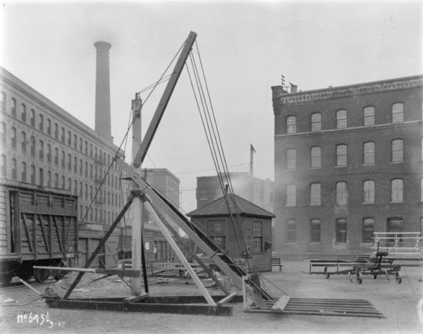 Hay stacker outdoors in a yard at McCormmick Works. There is a railroad car on tracks on the left. Factory buildings and a smokestack are in the background.