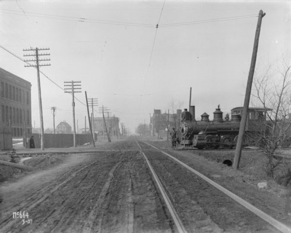 View down dirt road towards a railroad crossing. A man is standing near a fence on the left. A brick factory building is behind the fence, and more buildings are in the background. A man is standing on the front of a locomotive which is on the railroad tracks on the right side of the road, heading towards the factory.