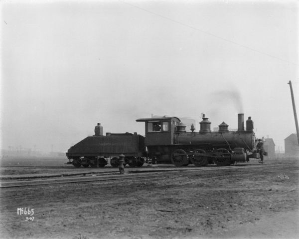 A man is standing on the front of a locomotive that is crossing a muddy road in the vicinity of Chicago.