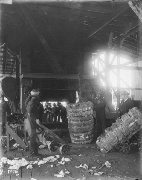 Men are standing inside a factory building unloading bales of manila or cotton. A man standing on the right is wearing a hat and suit.