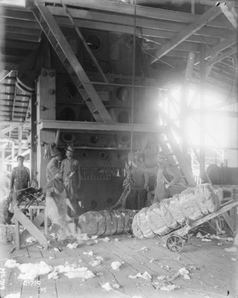 Men are standing inside a factory building unloading bales of manila or cotton.