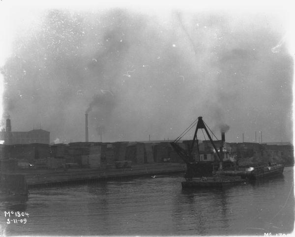 View across water towards ships in docks at McCormick Works. Lumber is in tall stacks along the wharf.