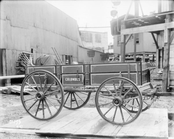 A Columbus Wagon is on a wooden platform outdoors. Factory buildings are in the background.