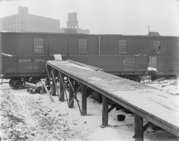 Dock over river with snow and ice flows. The dock leads to a railroad car. Factory buildings and water tanks are in the background.