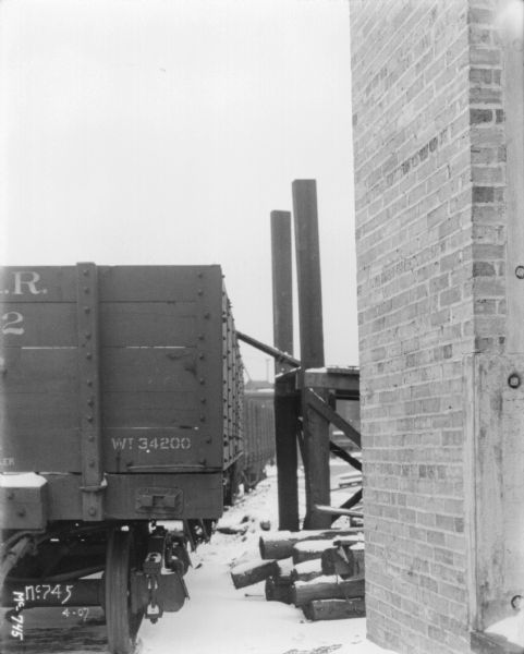 View towards the back of a railroad boxcar in snowy yard at McCormick Works. There is a brick wall of a factory building on the right, and a dock behind the building leads to the side of the boxcar.
