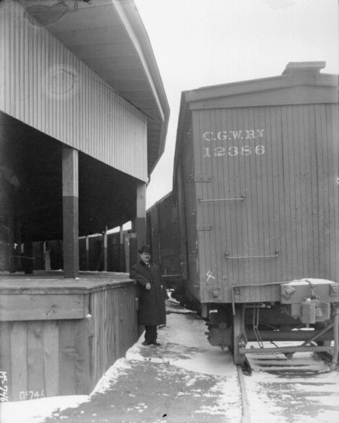 A man is standing near a covered platform of a building, with a row of boxcars alongside. Snow is on the ground.