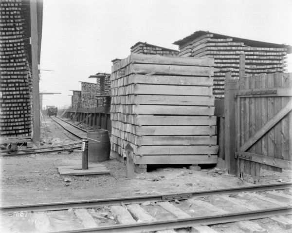 Lumber stacks at McCormick Works. There are railroad tracks in the foreground, and a gate and a fence on the right.
