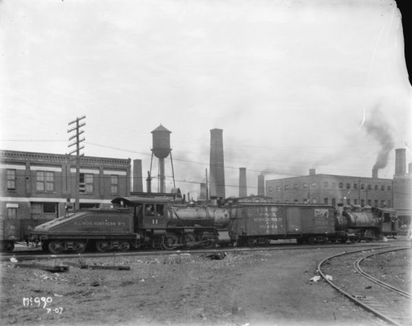 View towards the side of an Illinois Northern railroad train, with two engines, a coal car and a car with the front wall burst out. At McCormick Works. There are brick buildings, a water tower, and chimneys in the background.