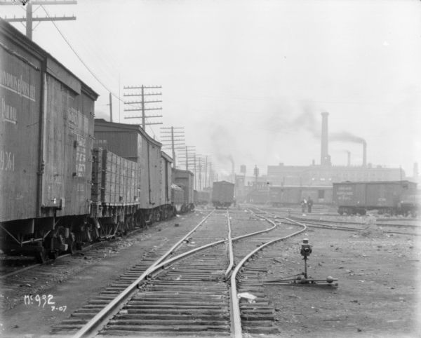 View down railroad tracks in a railroad yard at McCormick Works. There is a train along tracks on the left, and men are standing in the yard in the distance on the right.