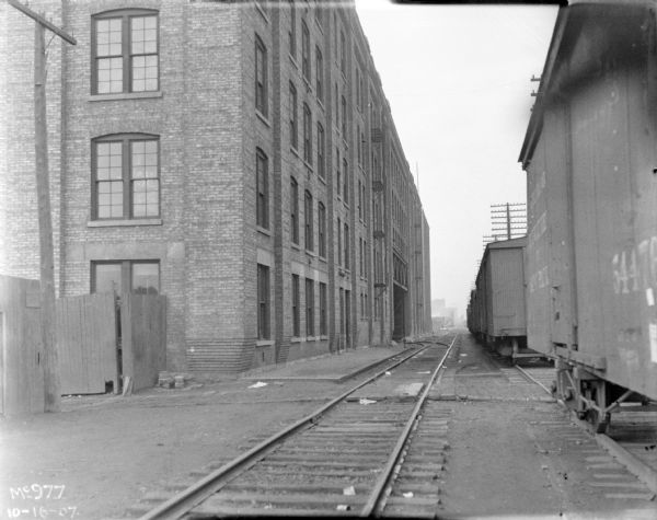 Railroad yard, with railroad cars on the right, and a fence and brick factory building on the left.