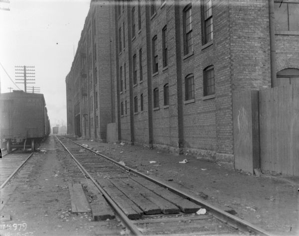 Railroad yard, with railroad cars on the left, and a fence and brick factory building on the right.