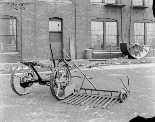 Rear view of a horse-drawn mower outdoors in a yard at McCormick Works. There is a brick factory building and a loading dock in the background.