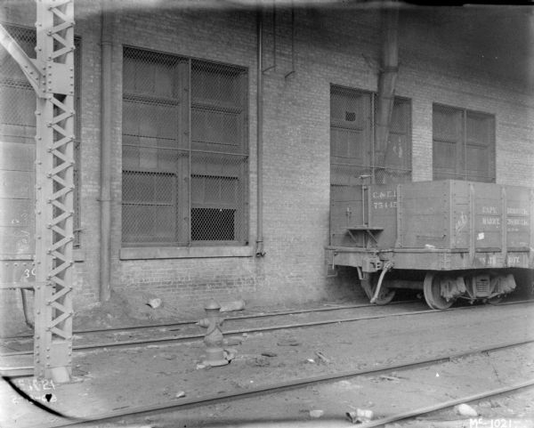 A railroad car is on railroad tracks under a roof alongside a brick factory building.